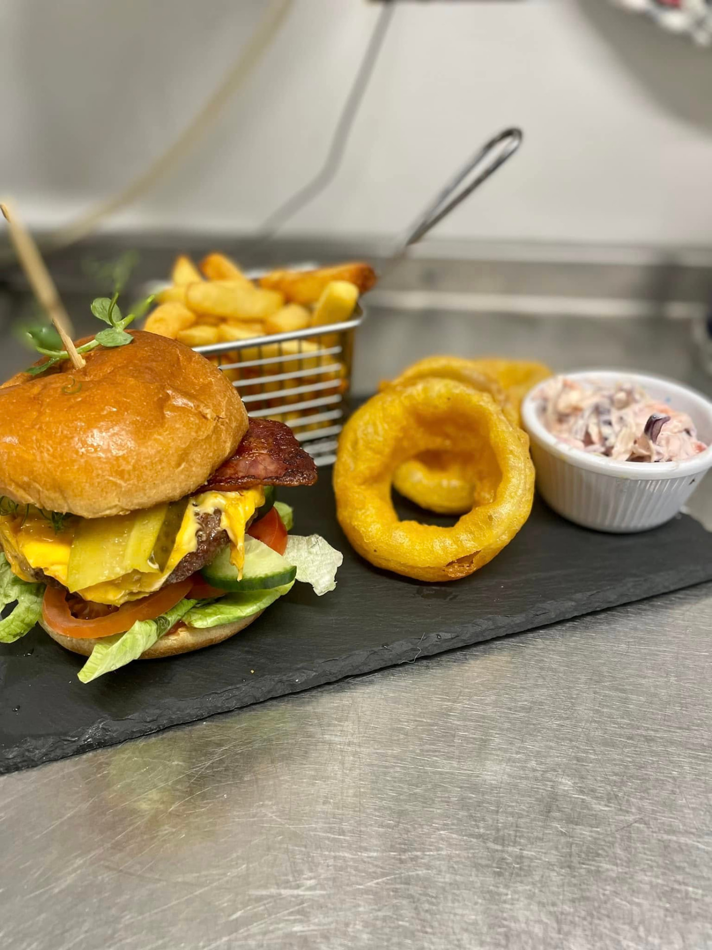 A bacon cheeseburger with chunky chips, onion rings and slaw on slate